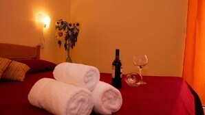 Ein ordentlich bezogenes Bett mit zwei aufgerollten Handtüchern, einer Weinflasche und einem Weinglas auf einer roten Tagesdecke.