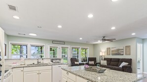 A spacious kitchen with white cabinetry, granite countertops, and a large island. There are two dark armchairs and a brown sofa in the adjacent living area.