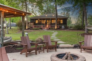 A cozy outdoor seating area with Adirondack chairs and a fire pit, a covered porch with string lights, and a house with a front porch and steps leading to the entrance.