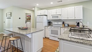A modern kitchen with white cabinets, granite countertops, a built-in oven, and a microwave. There are two bar stools and a welcome sign on the wall.