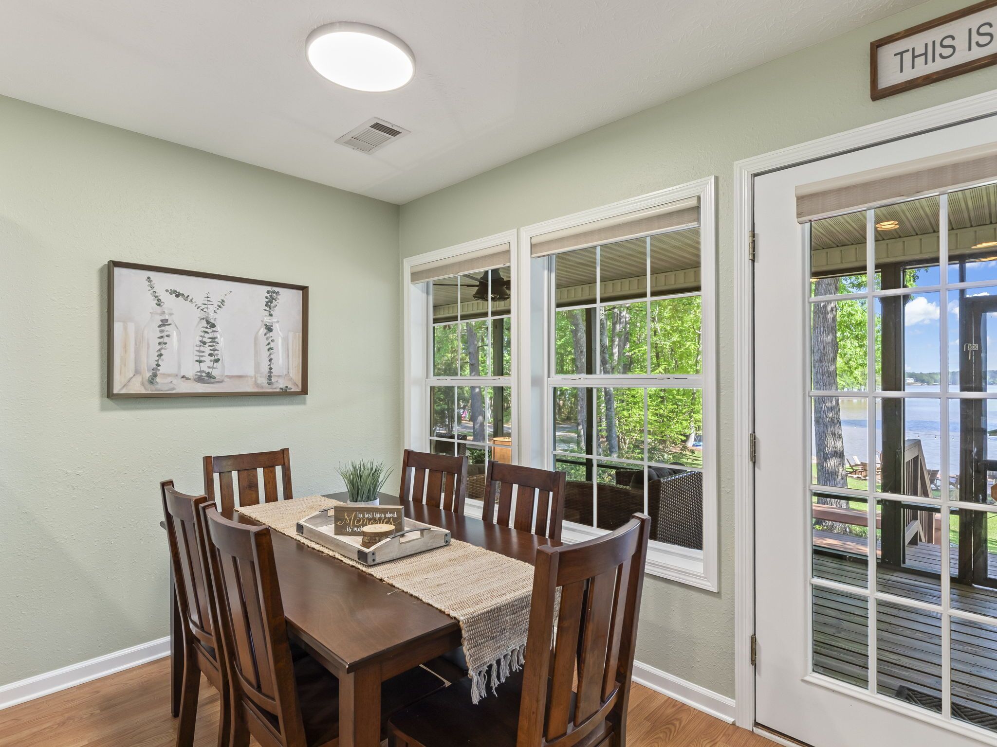 A dining area with a table and chairs, a framed artwork on the wall, and a view of a deck through large windows.