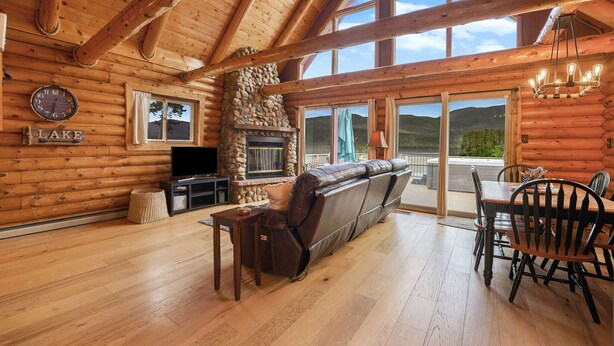 A log cabin interior with a stone fireplace, wooden floors, a sofa, a dining table with chairs, and a large window overlooking a mountain. - Lakefront A-Frame on Shadow Mountain Lake, Hot Tub, Kayaks & RMNP Views (Grand Lake)