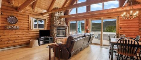 A log cabin interior with a stone fireplace, wooden floors, a sofa, a dining table with chairs, and a large window overlooking a mountain.