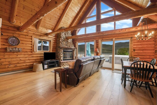 A log cabin interior with a stone fireplace, wooden floors, a sofa, a dining table with chairs, and a large window overlooking a mountain. - Lakefront A-Frame on Shadow Mountain Lake, Hot Tub, Kayaks & RMNP Views (Grand Lake)