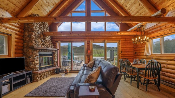 A log cabin interior with a stone fireplace, leather sofa, dining table, and chairs.