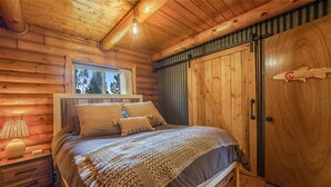 A log cabin bedroom with a bed, wooden walls, a window, a nightstand, and a door. - Lakefront A-Frame on Shadow Mountain Lake, Hot Tub, Kayaks & RMNP Views (Grand Lake)