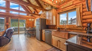 A log cabin interior with a kitchen, dining area, and a large window offering a mountain view.
