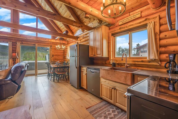 A log cabin interior with a kitchen, dining area, and a large window offering a mountain view.