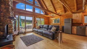 A log cabin interior with a stone fireplace, leather sofa, wooden floors, and a kitchen area.