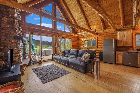 A log cabin interior with a stone fireplace, leather sofa, wooden floors, and a kitchen area. - Lakefront A-Frame on Shadow Mountain Lake, Hot Tub, Kayaks & RMNP Views (Grand Lake)