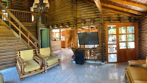 A log cabin interior with a staircase, seating area, television, and a kitchen in the background.