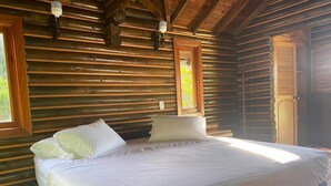A wooden cabin bedroom with a large bed, two wall-mounted lamps, and a window with a view of greenery.