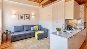 Una sala moderna con un sofá gris, una mesita auxiliar y una cocina con armarios blancos y encimera de mármol.
