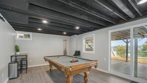 Une table de billard dans une pièce dotée d’un plancher en bois, d’un plafond avec des poutres apparentes et d’une vue sur un lac, visible à travers de grandes fenêtres.