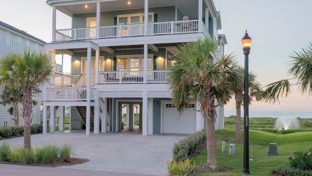 A multi-story building with balconies, a covered entrance, and a well-maintained lawn with palm trees. - Pointe West Beach Custom Home, Beach to Bay views, PW Pool/Private Beach (Galveston)