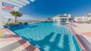 A swimming pool with a lifeguard warning sign, surrounded by resort buildings and palm trees. - Pointe West Beach Custom Home, Beach to Bay views, PW Pool/Private Beach (Galveston)