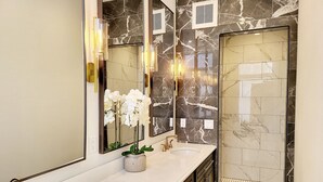 Un baño moderno con salpicadero de mármol, un espejo grande, un lavabo blanco, un grifo dorado y una zona de ducha.