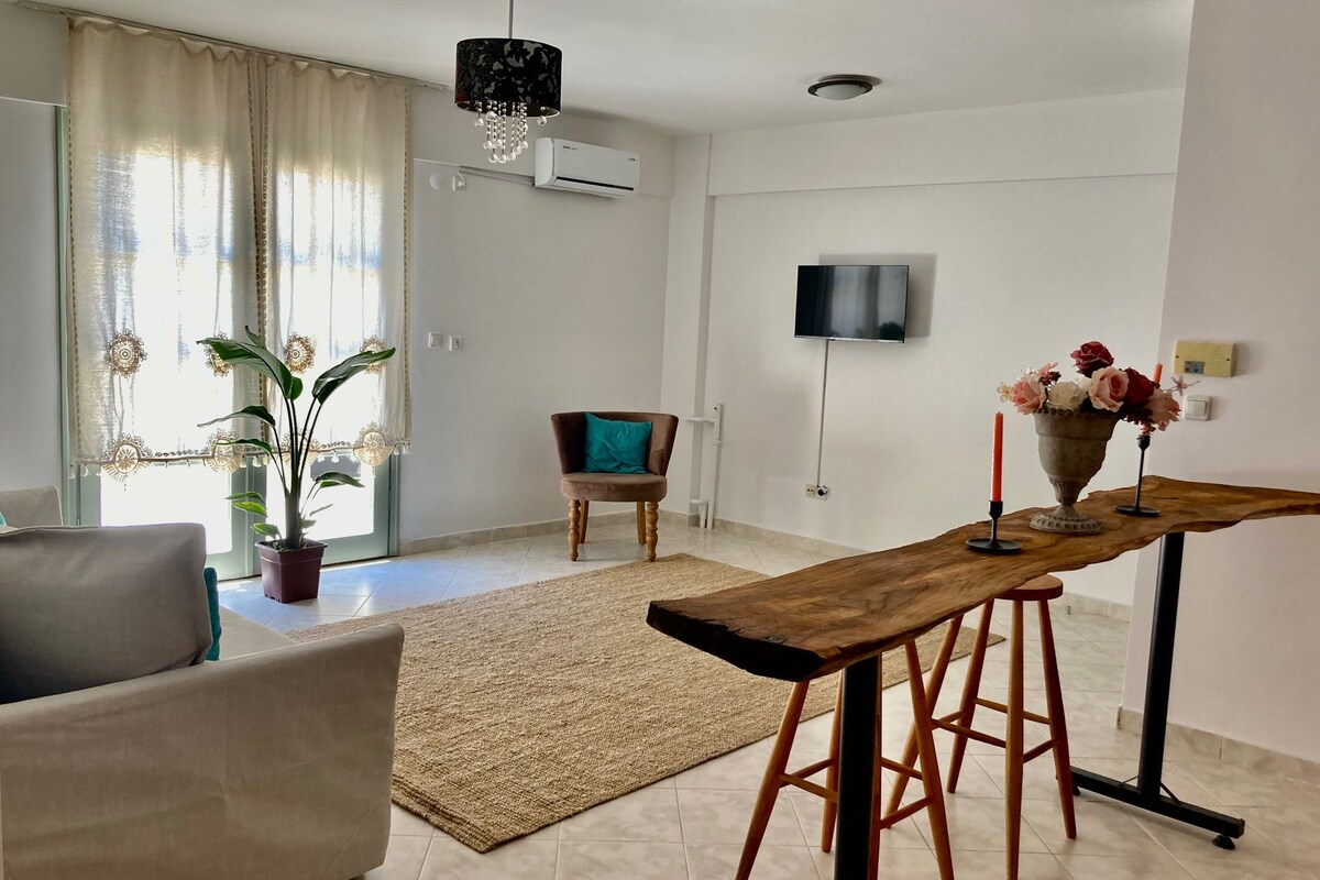 A modern living room with a sofa, a wooden table with stools, a potted plant, a TV mounted on the wall, and a ceiling fan.