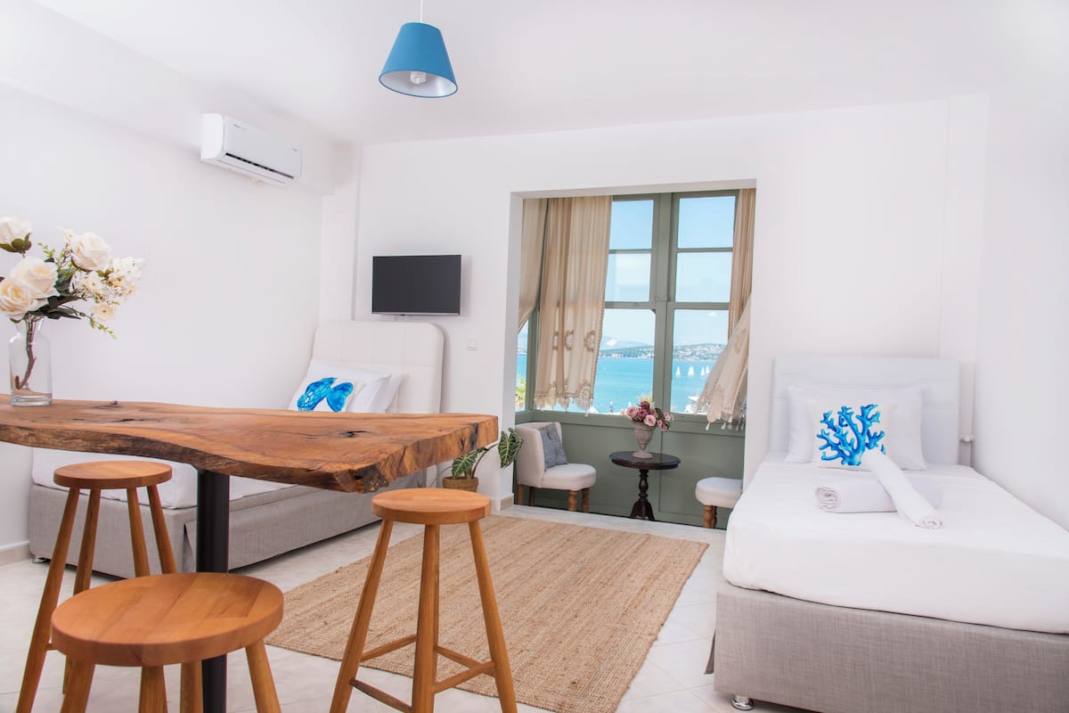 Una habitación de hotel moderna con una mesa de comedor de madera, dos taburetes de madera, un sofá, una cama, un televisor y vista al mar.