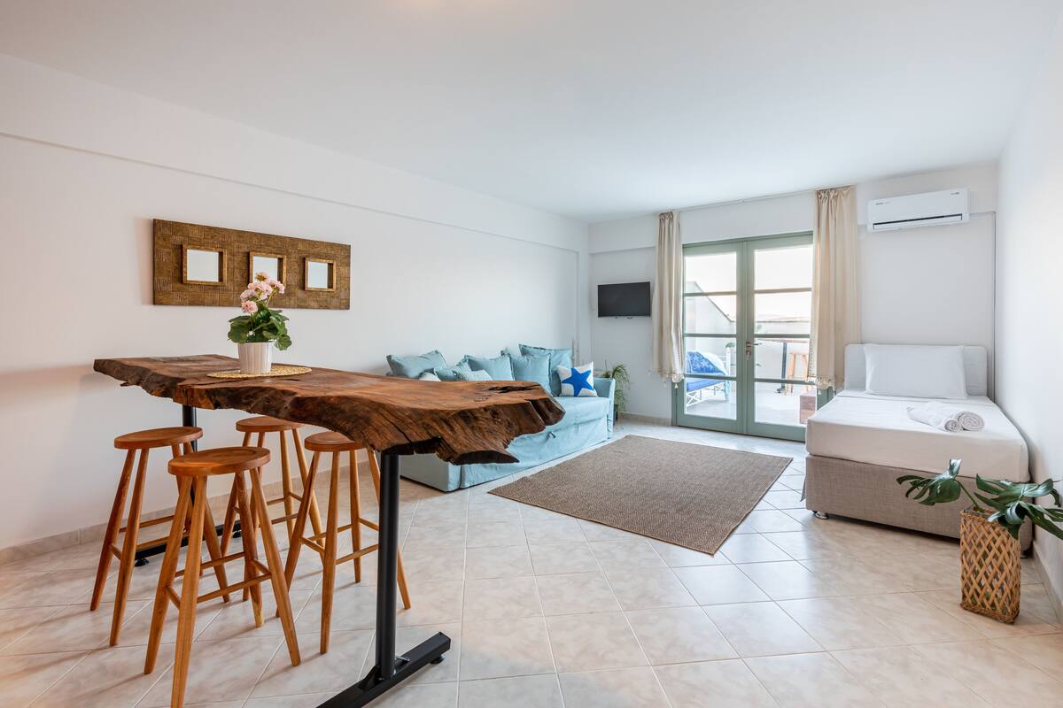 Una sala de estar moderna con una mesa de comedor de madera, taburetes de bar, un sofá con cojines azules, un ventanal grande y una cama con colchón blanco.