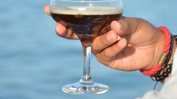 Eine Hand hält ein Glas dunkle Flüssigkeit, möglicherweise ein Getränk, im Hintergrund ist ein Gewässer und ein Boot zu sehen.