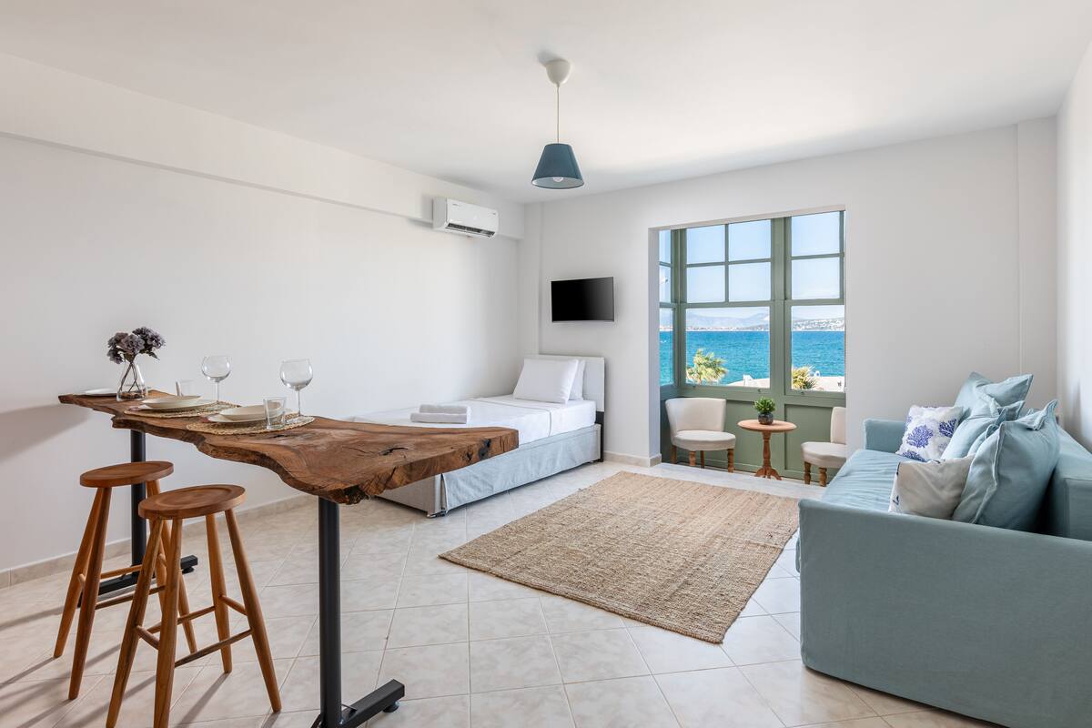 Una habitación moderna con cama, sofá, zona de comedor con taburetes, televisor y un amplio ventanal con vistas al mar.