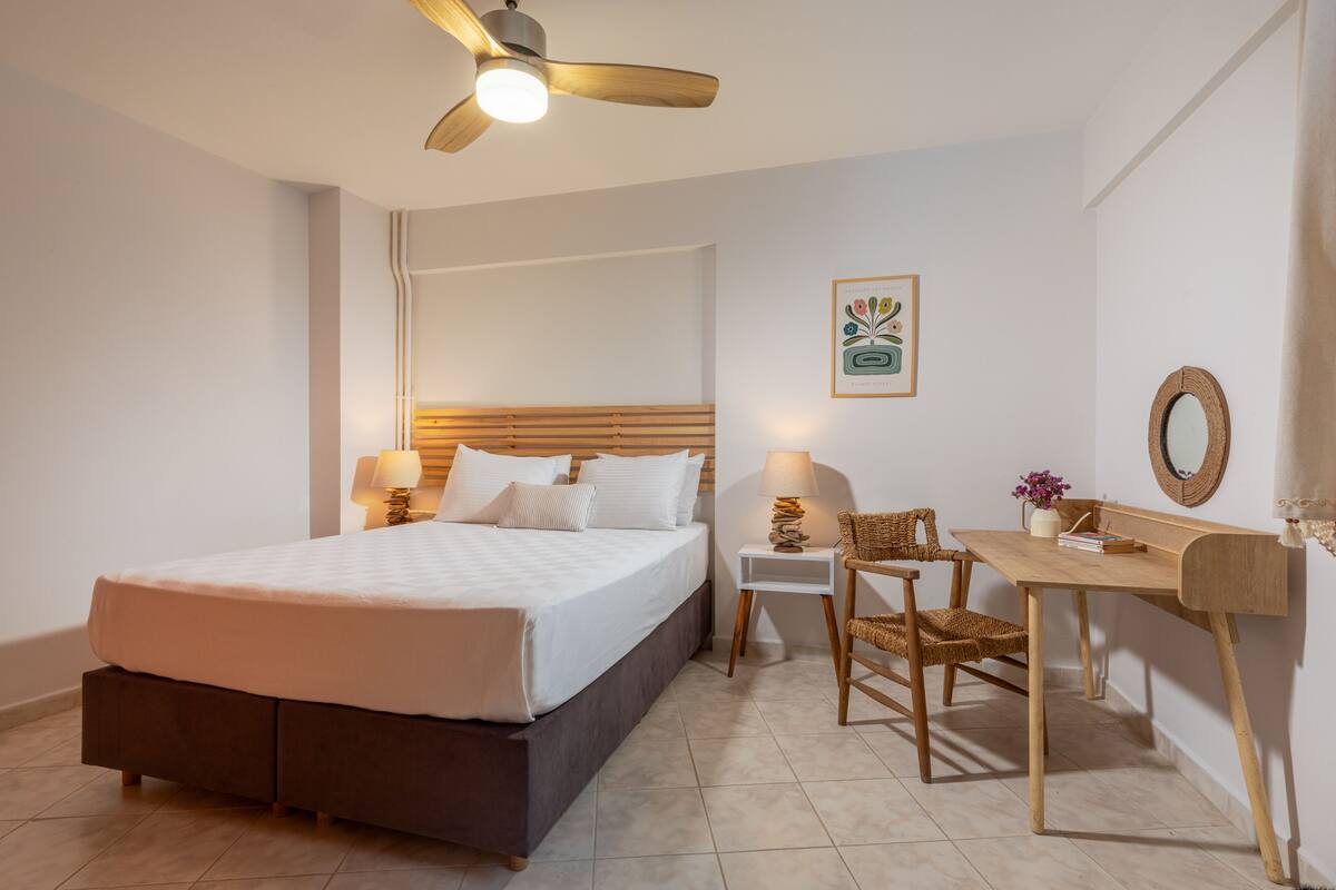 Un dormitorio con cama, mesitas de noche, escritorio, silla, ventilador de techo y espejo en la pared.