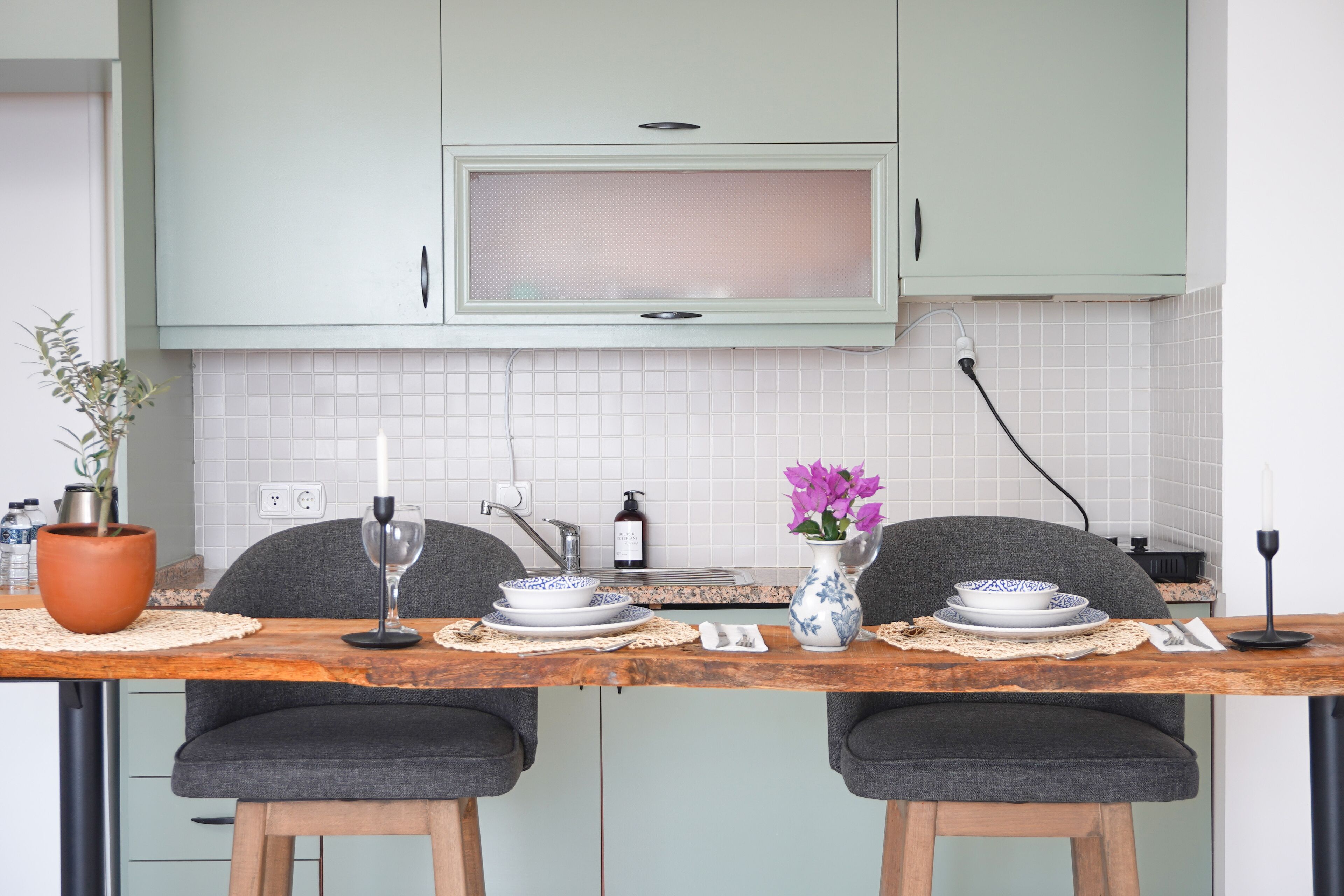 Une cuisine avec des meubles de couleur vert clair, un dosseret de carrelage blanc et une table en bois mise pour deux personnes, avec des assiettes, des verres et un vase rempli de fleurs.