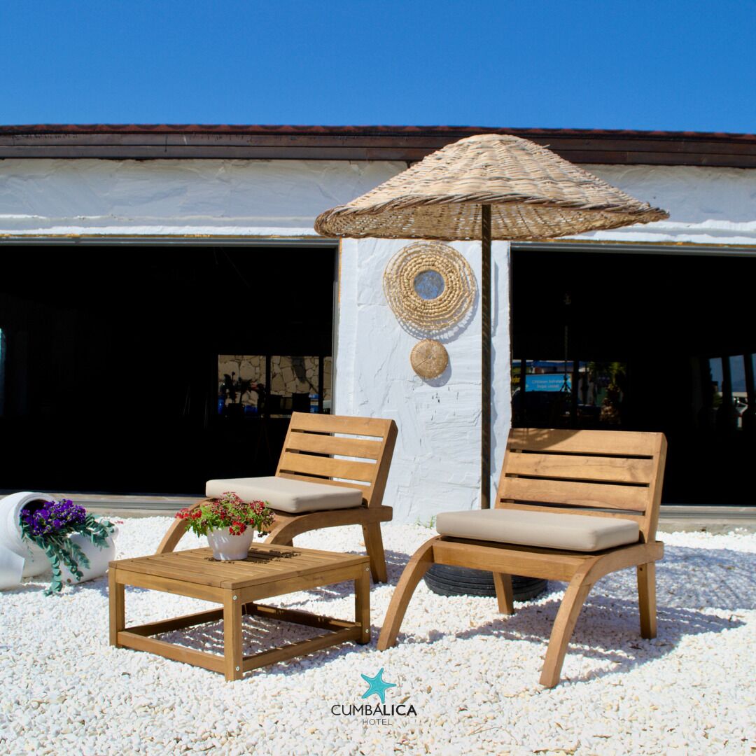 Two wooden lounge chairs with cushions, a small wooden table with a vase of flowers, and a woven umbrella. The setting includes a white wall with decorative woven items and a clear blue sky.