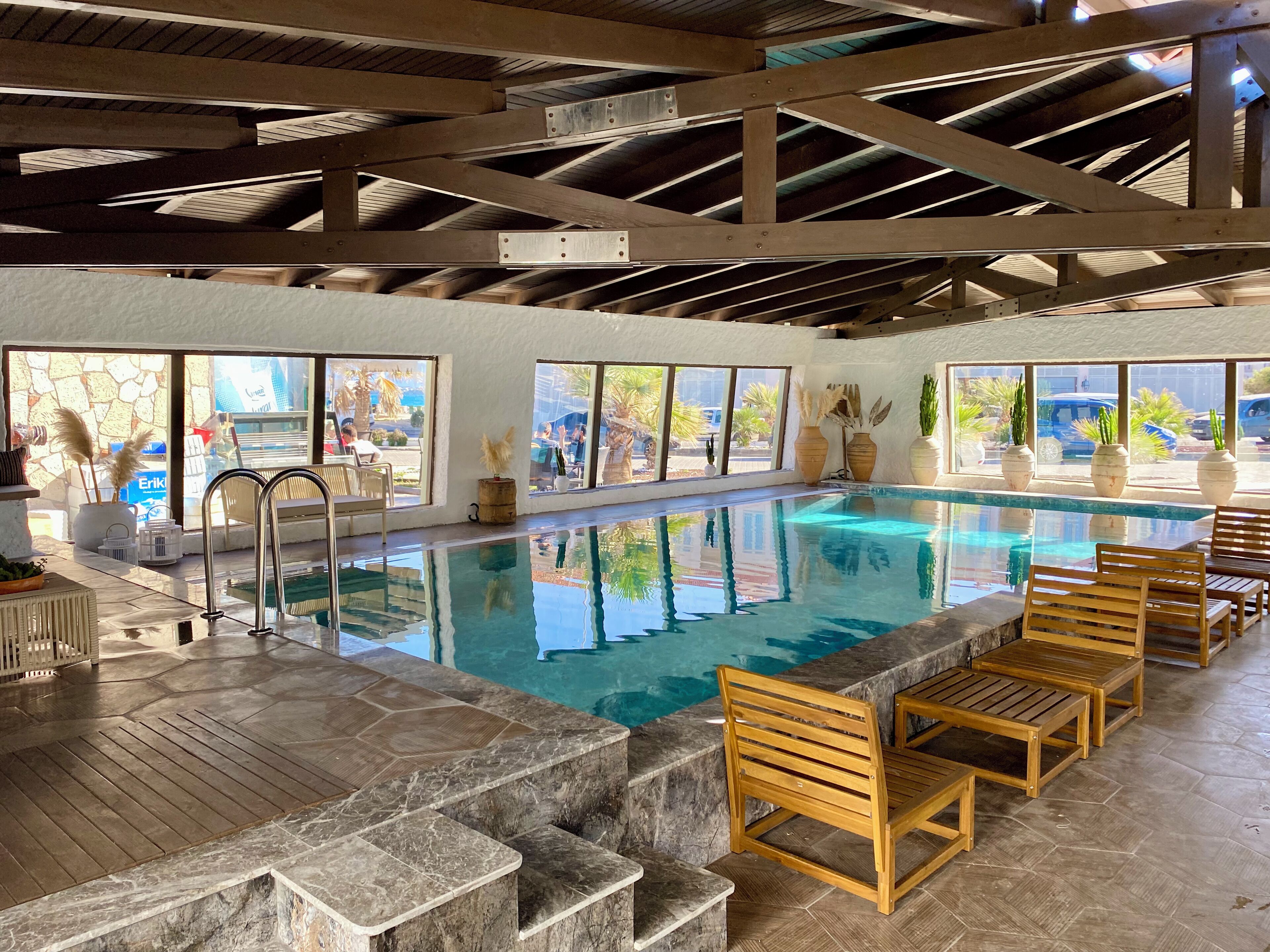 An indoor swimming pool with a view of the outside, wooden lounge chairs, and a stone-tiled floor.