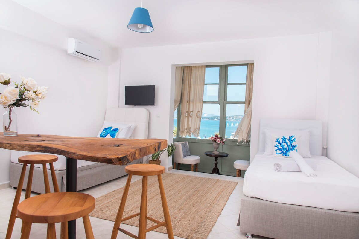 A modern hotel room with a wooden dining table, two wooden stools, a sofa, a bed, a TV, and a view of the sea.