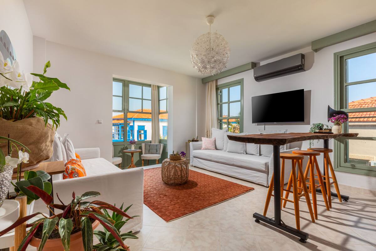 Una sala de estar moderna con un sofá, una mesa de centro y un televisor. Hay ventanas grandes y se ve una vista de edificios.