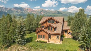 A two-story log cabin with a stone foundation, surrounded by a well-maintained lawn and tall trees, set against a mountainous backdrop. - Goldlink Lodge (Crested Butte)