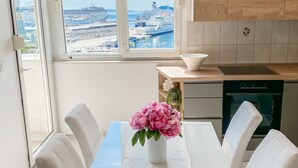 Une cuisine moderne avec un coin repas, de grandes fenêtres offrant une vue sur un navire de croisière, et un vase de fleurs sur la table.