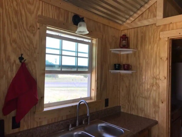 Interior de una cabaña de madera con cocina, que incluye un lavamanos, ventana y estantes.