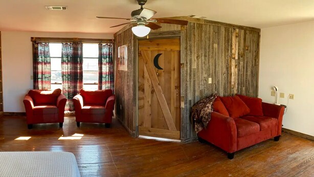 Une pièce avec un plancher en bois, un canapé rouge, deux fauteuils rouges, un ventilateur de plafond et une porte en bois ornée d’une découpe en forme de croissant de lune.