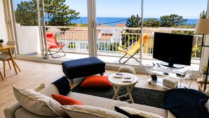 A modern living room with a sofa, ottoman, and chairs, a TV, and a balcony with a view of the sea. - Le Colombier - Ocean View (Les Sables-d´Olonne)