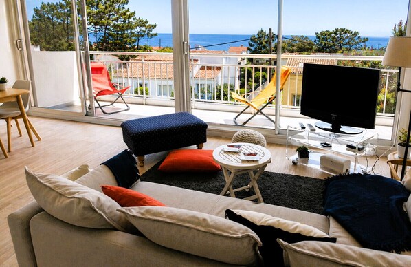 Un soggiorno moderno con divano, poggiapiedi e poltrone, televisione e un balcone vista mare.