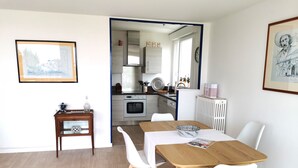 A modern kitchen with stainless steel appliances, a dining area with a wooden table and white chairs, and a small side table with decorative items. - Le Colombier - Ocean View (Les Sables-d´Olonne)