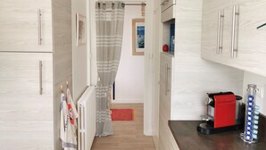 A narrow kitchen with light wood cabinets, a coffee machine, and a radiator. - Le Colombier - Ocean View (Les Sables-d´Olonne)