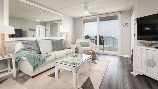 Ein modernes Wohnzimmer mit einem weißen Sofa, einem Glastisch und einem großen Spiegel. Ein Fernseher steht auf einer weißen Kommode und vom Balkon hat man einen Ausblick.