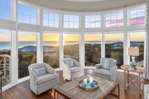 Una stanza con ampie finestre che offrono vista sul mare e sulle colline al tramonto. Sono presenti due poltrone e un tavolino in legno con oggetti decorativi.