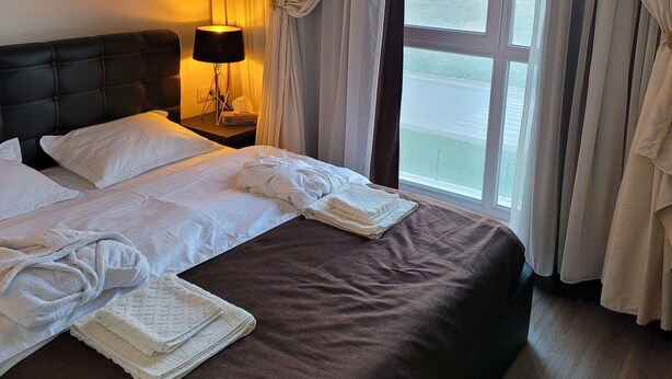 A hotel room with a large bed, a nightstand with a lamp, and a view of a body of water through the window. - Luxury Apartment in Tbilisi | 24/7 Security | Prime Location (T'bilisi)