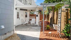 Une maison à deux étages dotée d’un patio couvert, d’un petit jardin avec des plantes et un arbre, ainsi que d’une clôture blanche.