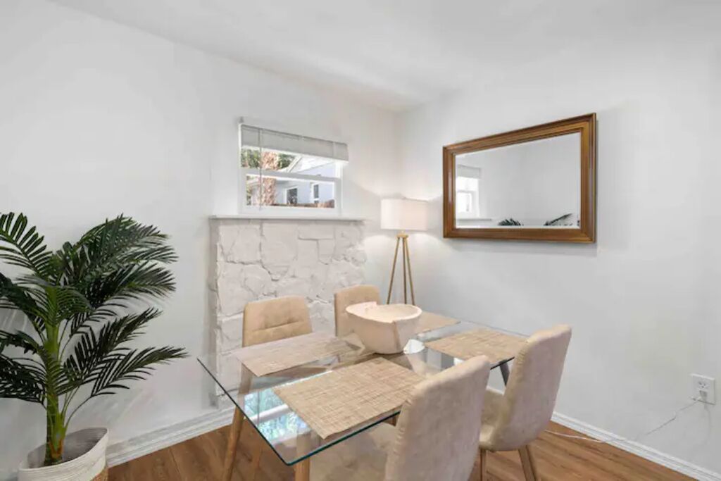 A dining area with a glass table, four chairs, a potted plant, a stone wall, a mirror, and a lamp.
