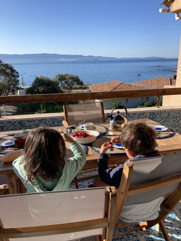 A dining table with a view of the sea, a bowl of strawberries, and a teapot. - Duplex terrace with sea view 3 minutes' walk from the most beautiful beaches (Ajaccio)