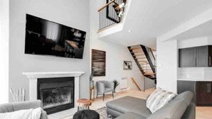 Una sala de estar moderna con chimenea, un televisor de pantalla plana grande, un sofá gris y una escalera que lleva a un nivel superior.