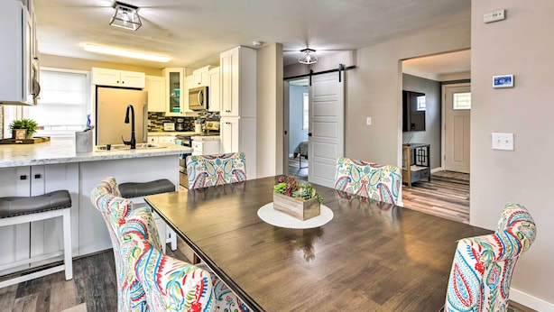 Une cuisine moderne avec des meubles blancs, un îlot central et un coin repas comprenant une table en bois et des chaises aux motifs colorés.