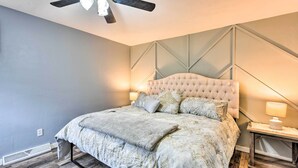 Une chambre à coucher avec une tête de lit capitonnée, un lit recouvert d’une couette à motifs, un plancher en bois et un ventilateur de plafond.