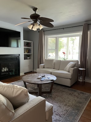 A living room with a sofa, a coffee table, a fireplace, a bookshelf, and a large window with curtains. - Private and Quiet Beach House! (Emerald Isle)